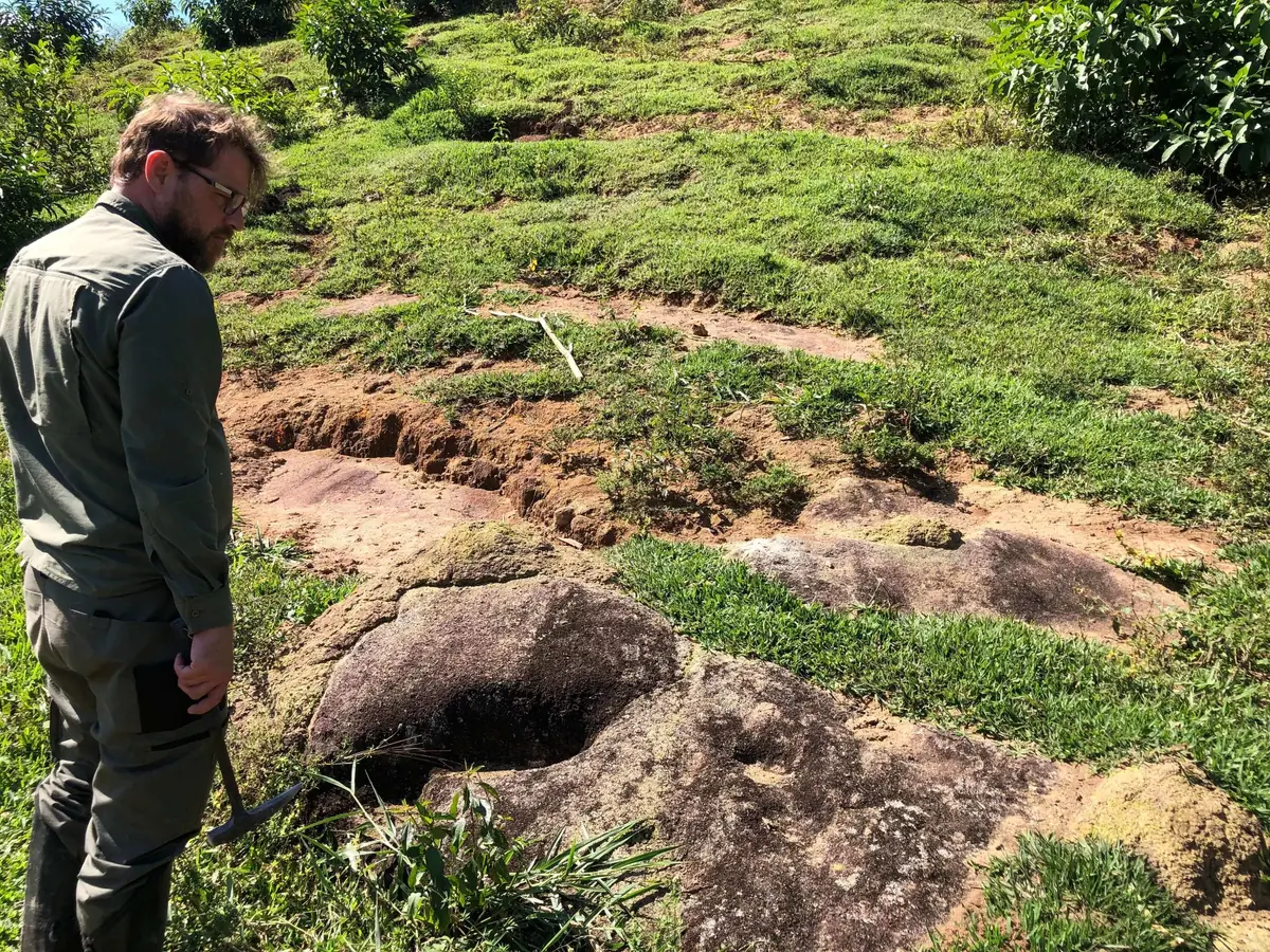 Field geologist examining weathered granite with surface cavities and vegetation cover.