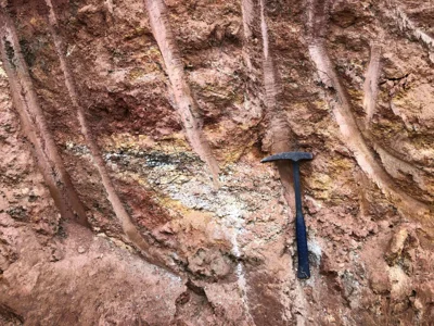 Pegmatitic vein exposure with quartz-feldspar bands and iron-stained weathering, hammer for scale.