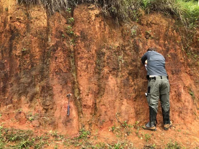 Geologist sampling reddish-brown residual soil over weathered schist
