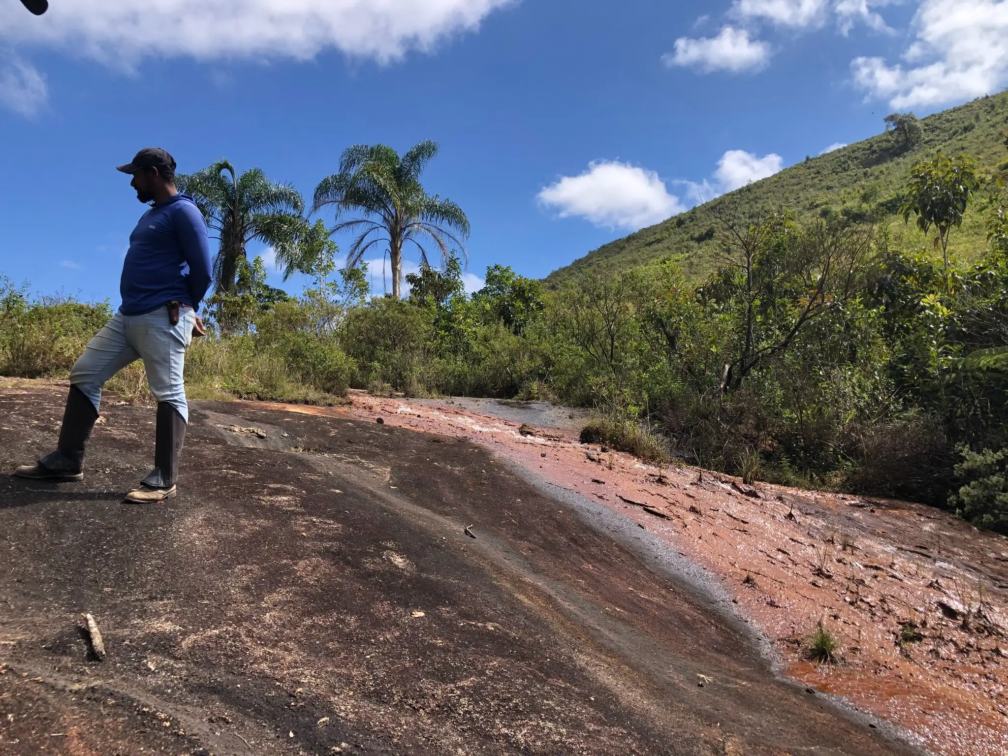 Field geologist standing beside a shallow oxidized drainage channel cutting through exposed granitic bedrock.