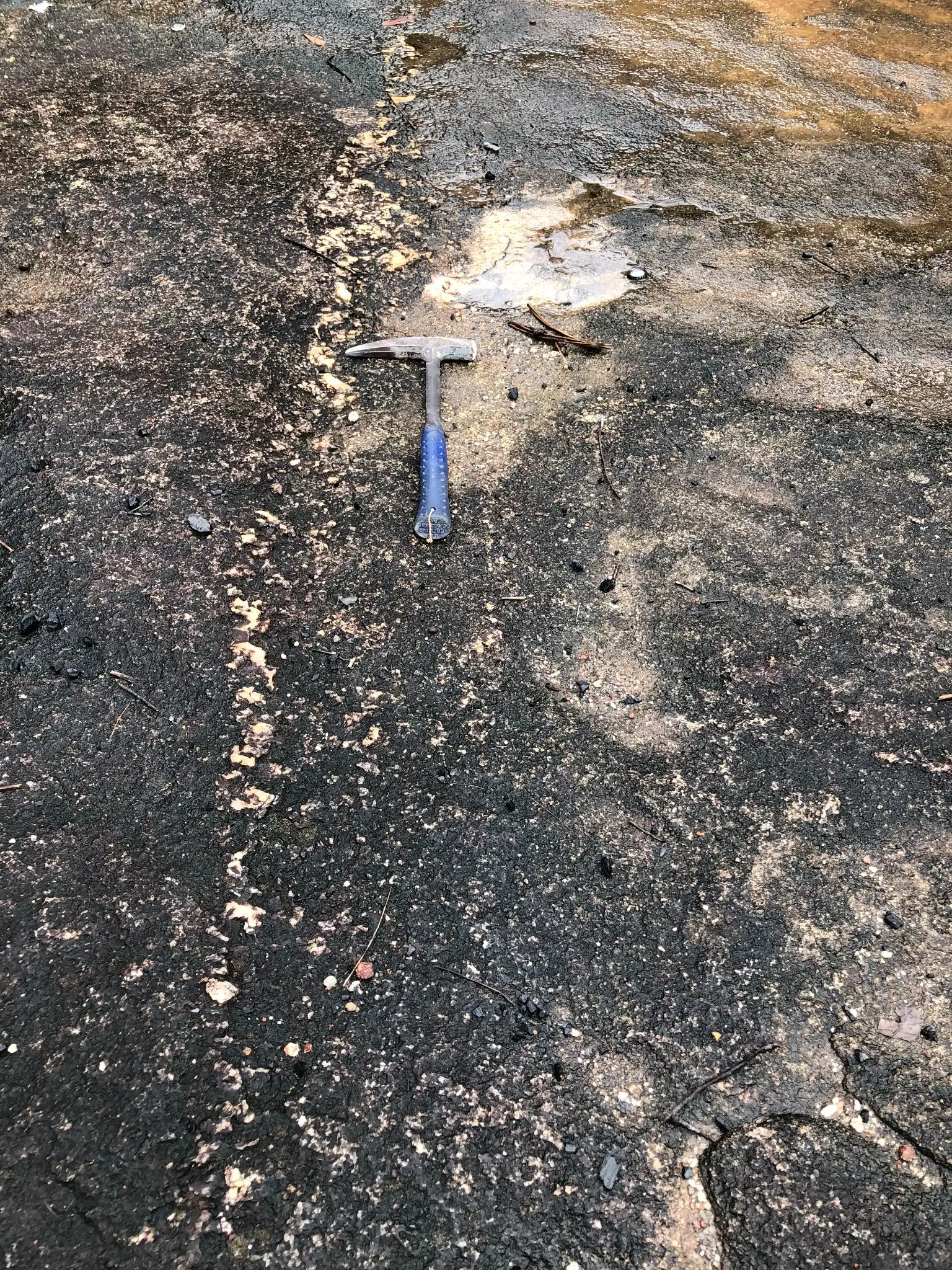 Rock hammer on a darkened granitic outcrop showing fracture-controlled oxidation and light surface water flow.