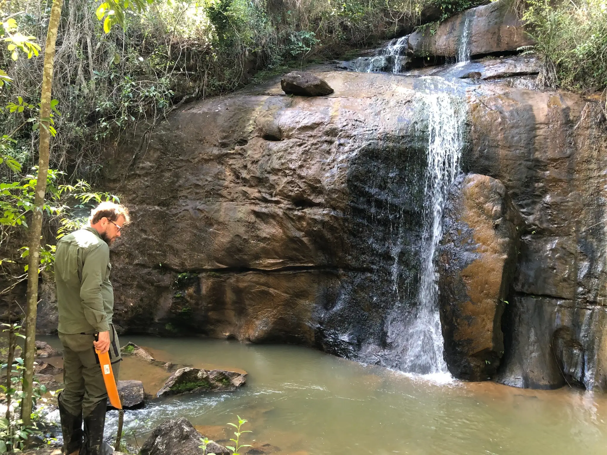 Field geologist collecting a stream sediment sample below a waterfall cutting through granitic bedrock.