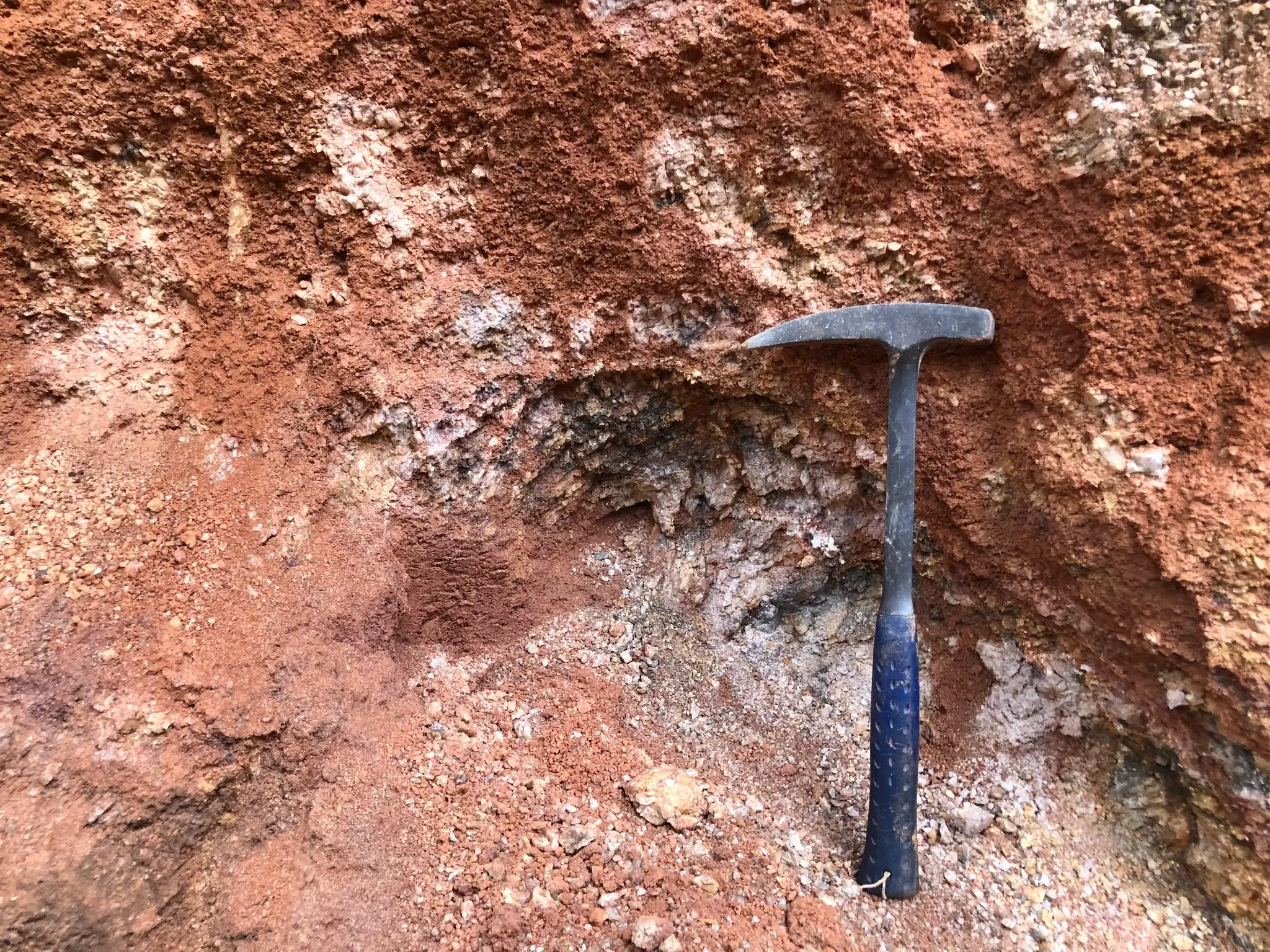 Weathered exposure with red-brown saprolite, pale clay-rich material, and quartz fragments.