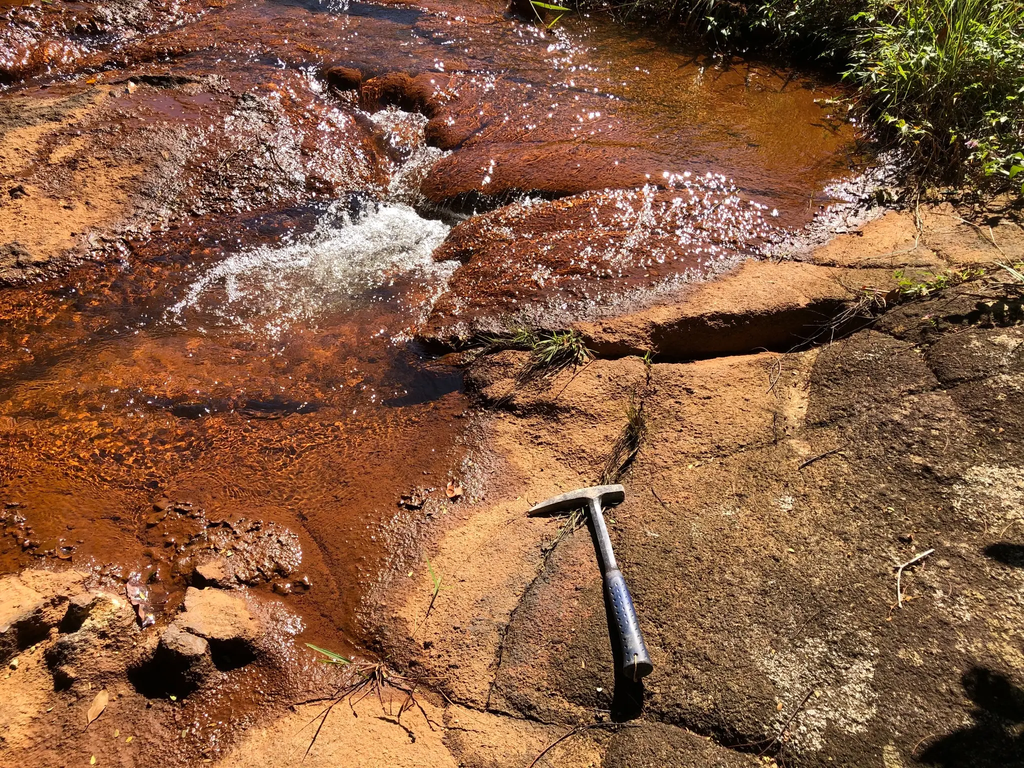 Stream channel over weathered granitic bedrock with iron staining and hammer for scale.