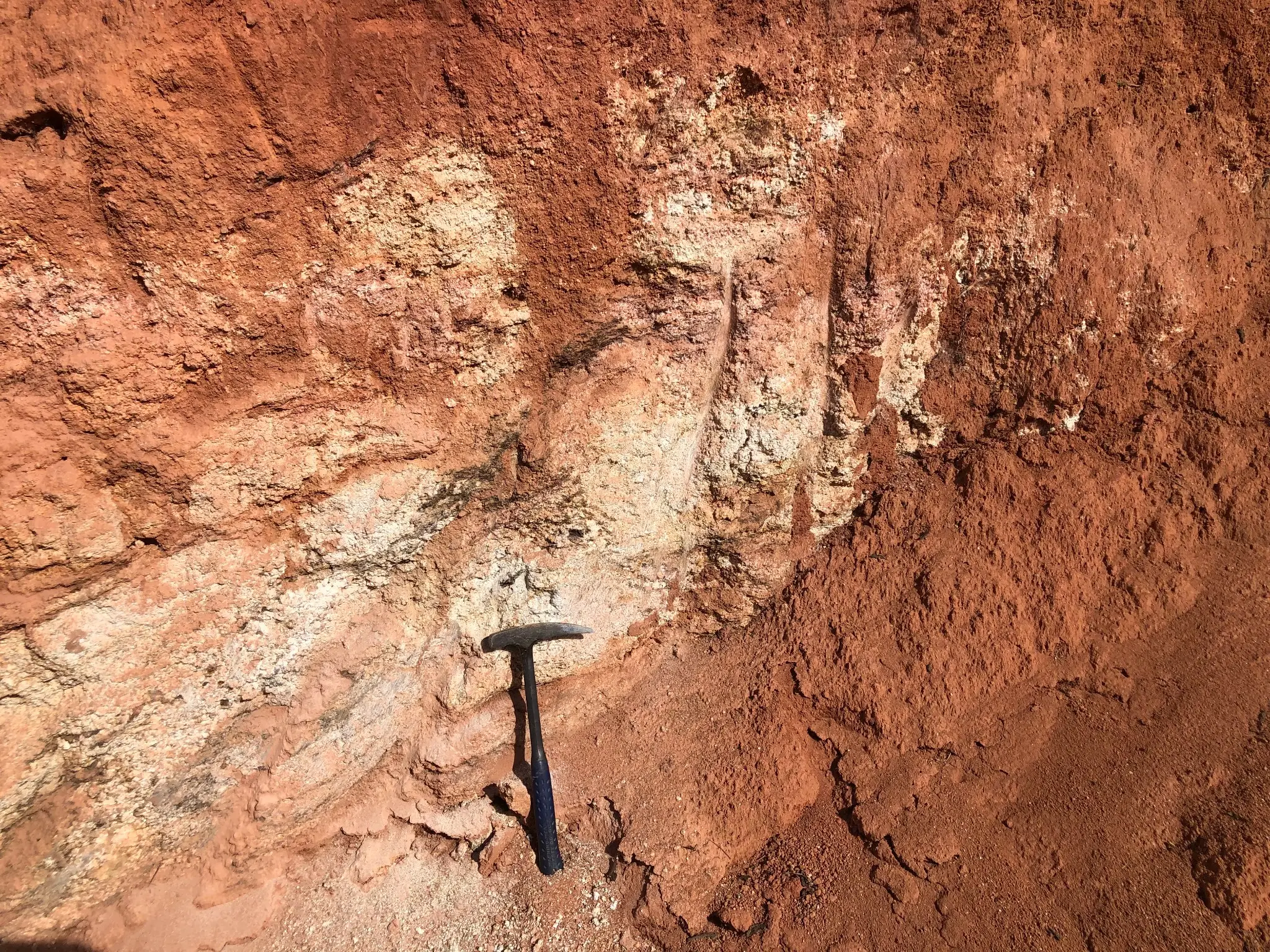 Red-weathered granitic exposure with quartz-rich veinlets and hammer for scale.