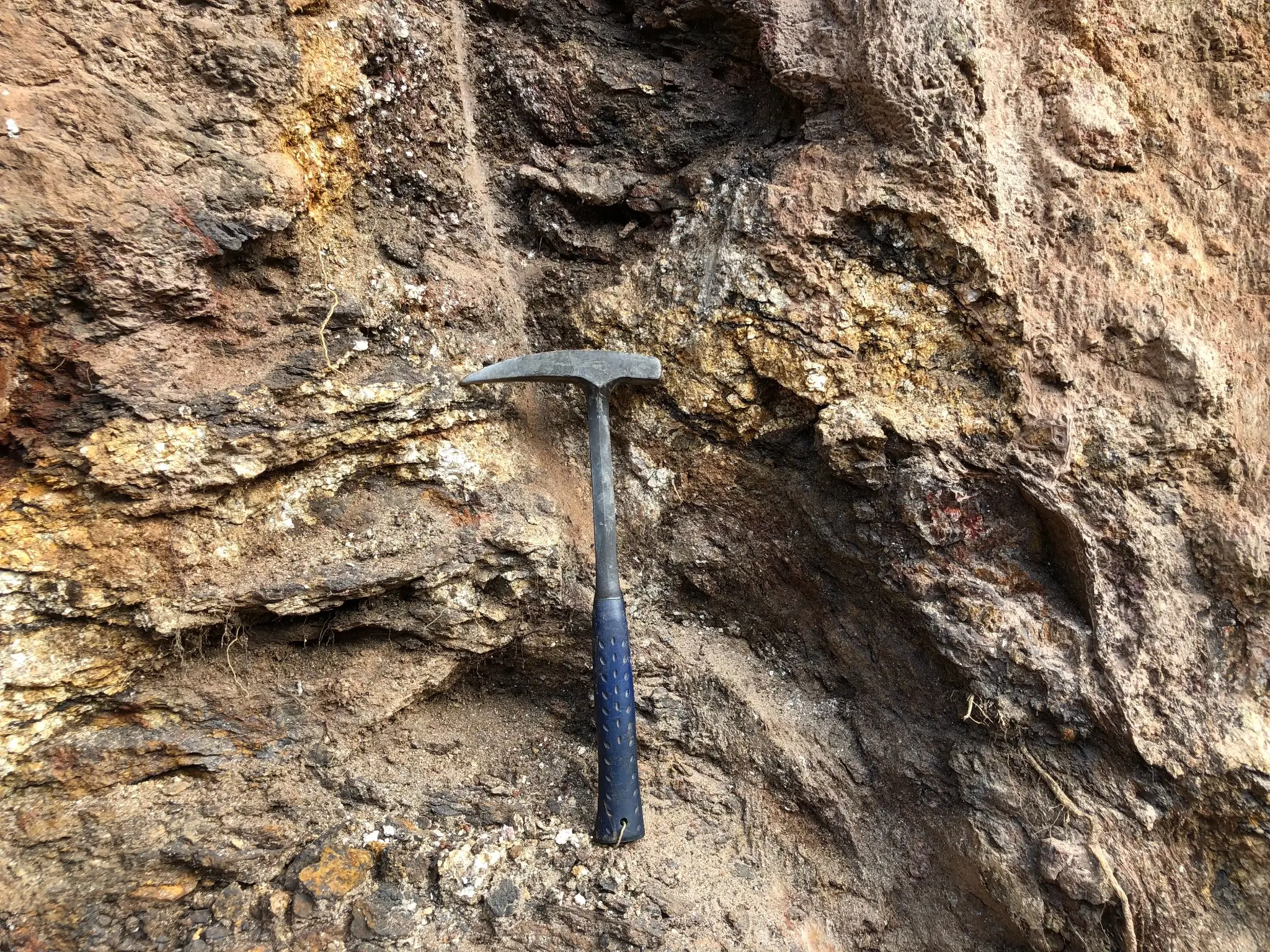 Field hammer beside an iron-stained weathered outcrop with clay-rich zones.